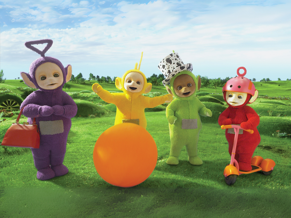 Laa-Laa, the yellow Teletubby, and Po, the red Teletubby standing in the grass. Po is holding a large orange ball.