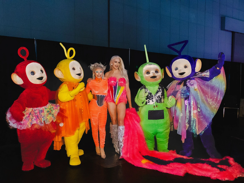 The Teletubbies pose against a pink DragCon background with a drag queen in a blue and pink dress.
