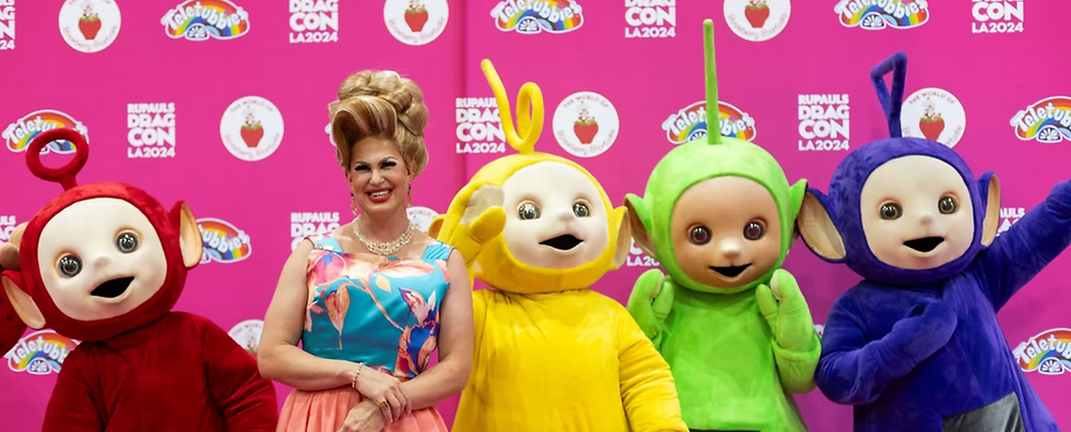 The Teletubbies pose against a pink DragCon background with a drag queen in a blue and pink dress.